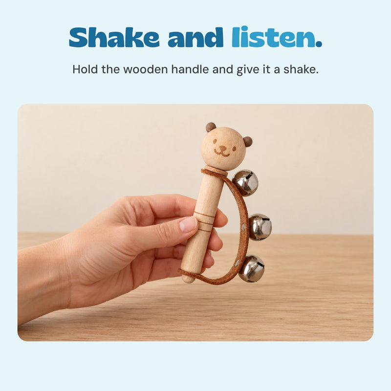 Adult hand holding the wooden bear bell rattle, with its curved brown strap and three silver bells visible.