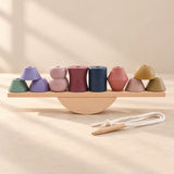 Muted geometric wooden pieces arranged on a straight balance plank and semicircular rocker, with the threading cord beside them.