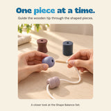 Two adult hands guide the wooden threading tip toward a lilac wooden shape, with a second shape already on the cotton cord.