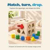 A hand turns a red wooden shape toward the cube, with colourful sorting pieces on the table.