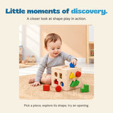 A child in a grey shirt holds a blue cylinder above the wooden sorting cube on a woven rug.