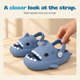 An adult hand holds the heel strap of a blue shark slide beside its matching shoe.