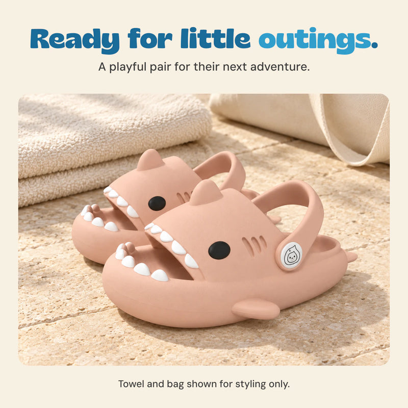 A pink pair of shark slides on a beige patio beside a styling towel and canvas bag. Towel and bag are not included.