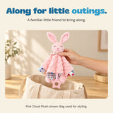 An adult hand holds the pink bunny comfort blanket above a cream canvas bag.