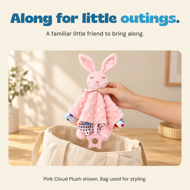 An adult hand holds the pink bunny comfort blanket above a cream canvas bag.