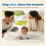 An awake baby lies on their tummy in the cream lounger while two grown-ups stay close by.