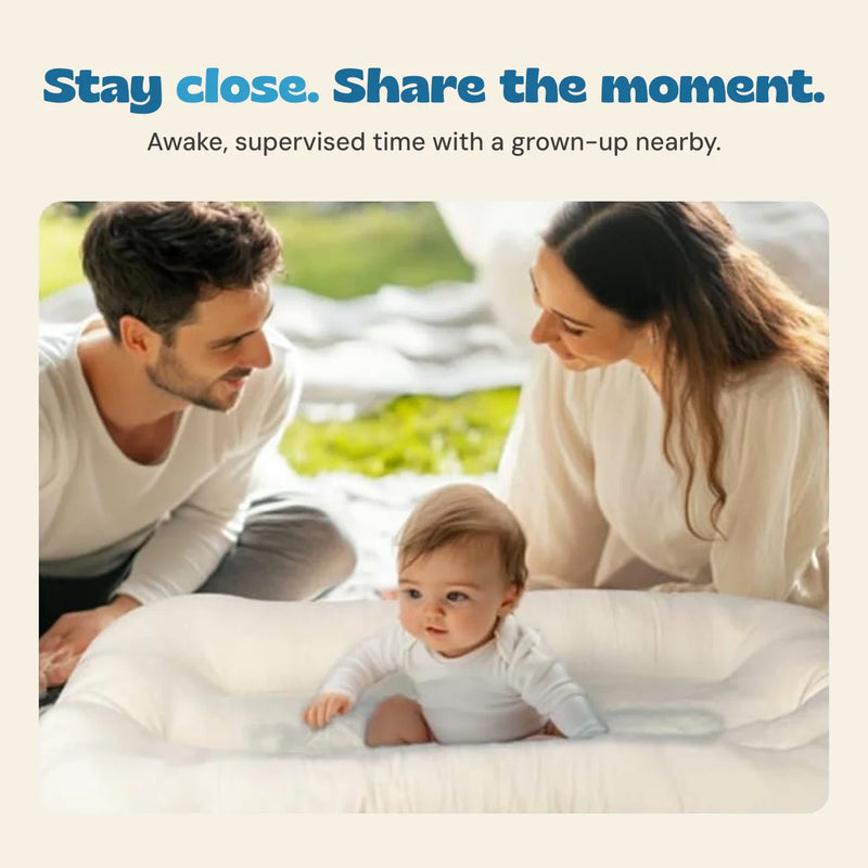 An awake baby lies on their tummy in the cream lounger while two grown-ups stay close by.