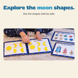 Hands arrange yellow moon shapes in an open Space quiet book, beside planet and rocket activity pages.