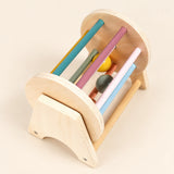 Wooden spinning drum with coloured bars and two balls in a natural wood stand on a pale beige background.