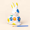Painted ceramic Easter bunny with blue, orange and yellow flowers beside a blue paintbrush on a warm beige background. Painted design shown as an activity example.