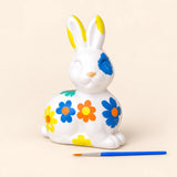 Painted ceramic Easter bunny with blue, orange and yellow flowers beside a blue paintbrush on a warm beige background. Painted design shown as an activity example.