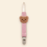 Pink fabric pacifier clip with brown teddy applique, cream loop and grey clasp on a pale warm beige background.