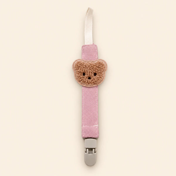 Pink fabric pacifier clip with brown teddy applique, cream loop and grey clasp on a pale warm beige background.