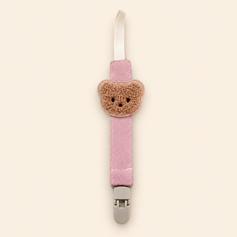 Pink fabric pacifier clip with brown teddy applique, cream loop and grey clasp on a pale warm beige background.