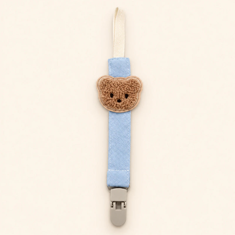 Light blue fabric pacifier clip with brown teddy applique, cream loop and grey clasp on a pale warm beige background.