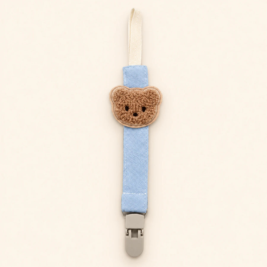 Light blue fabric pacifier clip with brown teddy applique, cream loop and grey clasp on a pale warm beige background.
