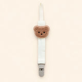 White fabric pacifier clip with brown teddy applique, cream loop and grey clasp on a pale warm beige background.