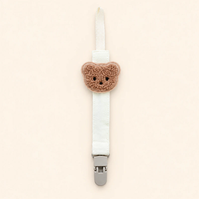 White fabric pacifier clip with brown teddy applique, cream loop and grey clasp on a pale warm beige background.