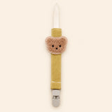 Mustard yellow fabric pacifier clip with brown teddy applique, cream loop and grey clasp on a pale warm beige background.