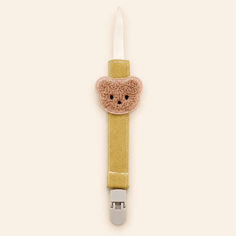 Mustard yellow fabric pacifier clip with brown teddy applique, cream loop and grey clasp on a pale warm beige background.