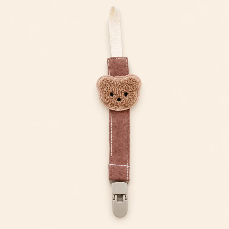 Dusty rose fabric pacifier clip with brown teddy applique, cream loop and grey clasp on a pale warm beige background.