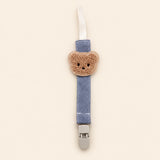 Slate blue fabric pacifier clip with brown teddy applique, cream loop and grey clasp on a pale warm beige background.