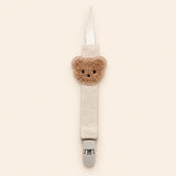 Cream fabric pacifier clip with brown teddy applique, cream loop and grey clasp on a pale warm beige background.