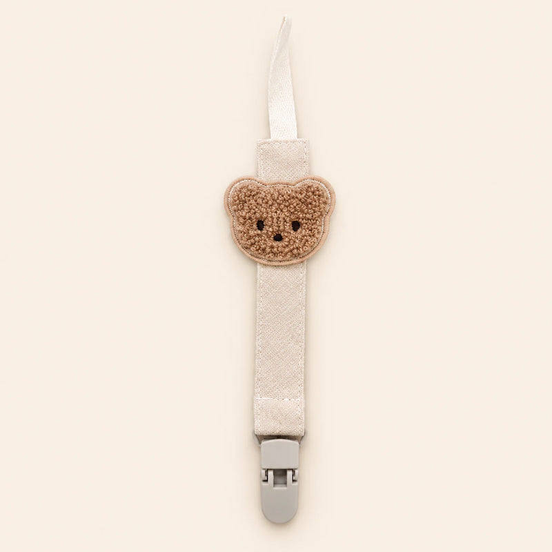 Cream fabric pacifier clip with brown teddy applique, cream loop and grey clasp on a pale warm beige background.
