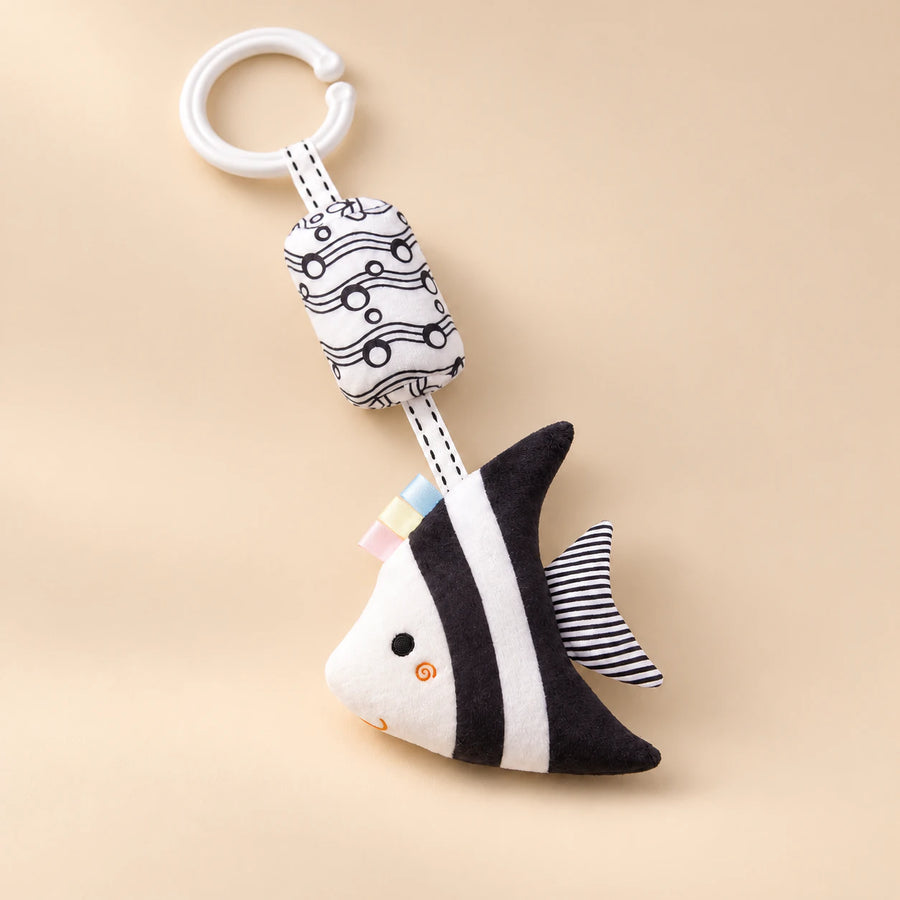 Black and white angelfish hanging rattle with its clip and patterned cushion on a sunlit beige tabletop.