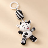Black and white cow hanging rattle with its clip and patterned cushion on a sunlit beige tabletop.
