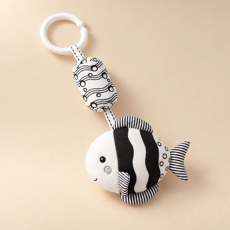 Black and white round fish hanging rattle with its clip and patterned cushion on a sunlit beige tabletop.