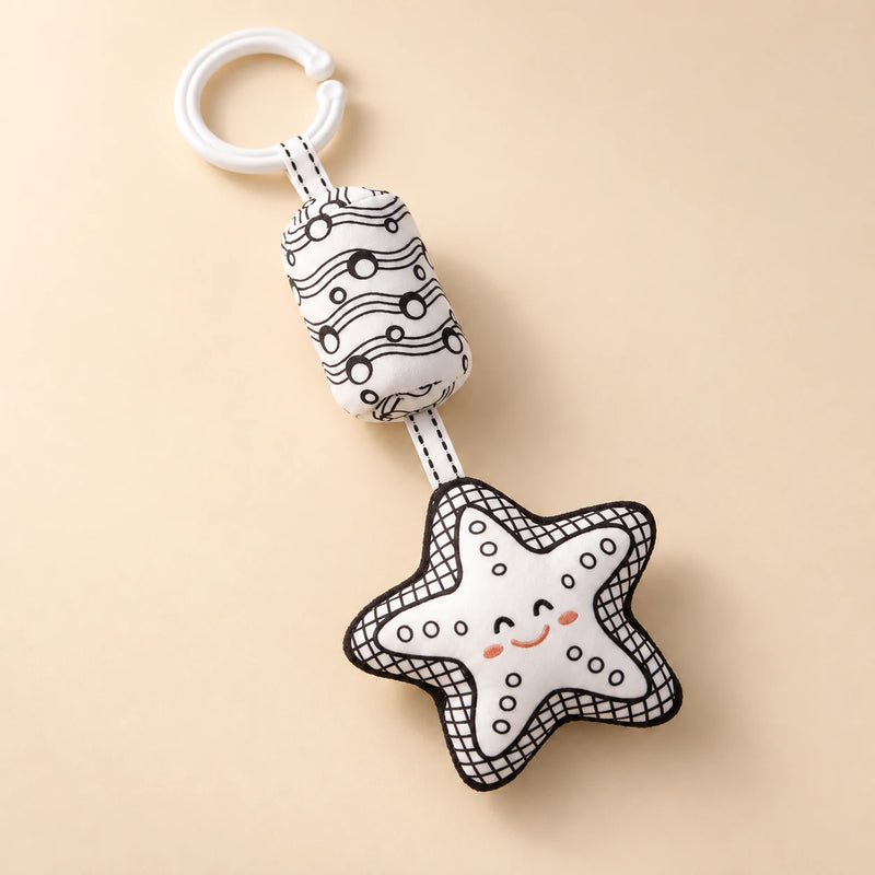 Black and white starfish hanging rattle with its clip and patterned cushion on a sunlit beige tabletop.