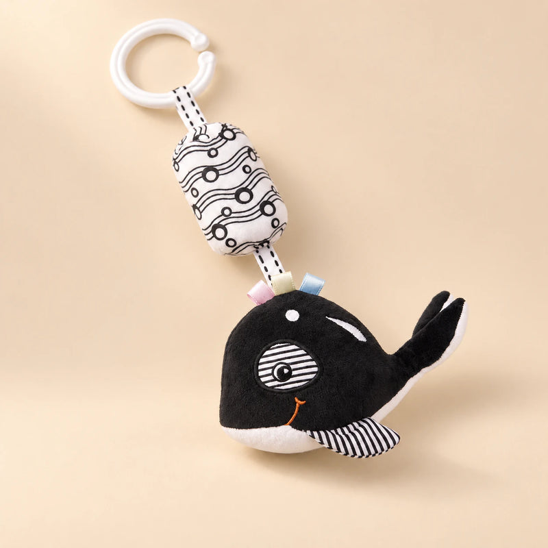 Black and white whale hanging rattle with its clip and patterned cushion on a sunlit beige tabletop.