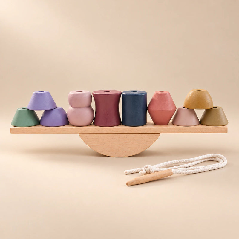 Muted geometric wooden pieces arranged on a straight balance plank and semicircular rocker, with the threading cord beside them.