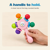 An adult hand holds the pink rattle handle while all five textured ends remain visible.