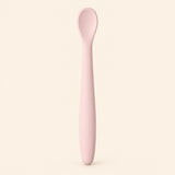 Blush silicone baby feeding spoon, showing its slim handle and shallow oval bowl on warm beige.