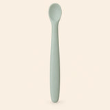 Sage silicone baby feeding spoon, showing its slim handle and shallow oval bowl on warm beige.