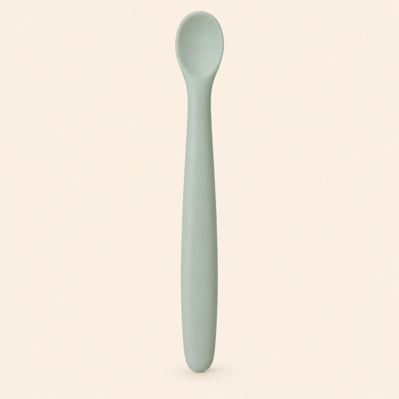 Sage silicone baby feeding spoon, showing its slim handle and shallow oval bowl on warm beige.