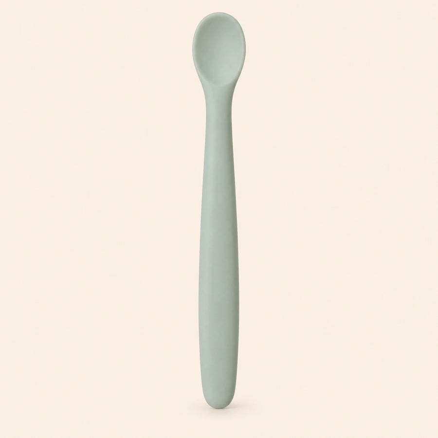Sage silicone baby feeding spoon, showing its slim handle and shallow oval bowl on warm beige.