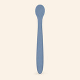 Denim silicone baby feeding spoon, showing its slim handle and shallow oval bowl on warm beige.
