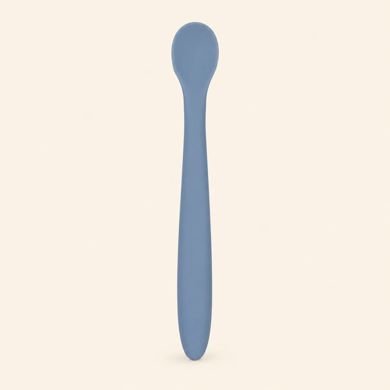 Denim silicone baby feeding spoon, showing its slim handle and shallow oval bowl on warm beige.