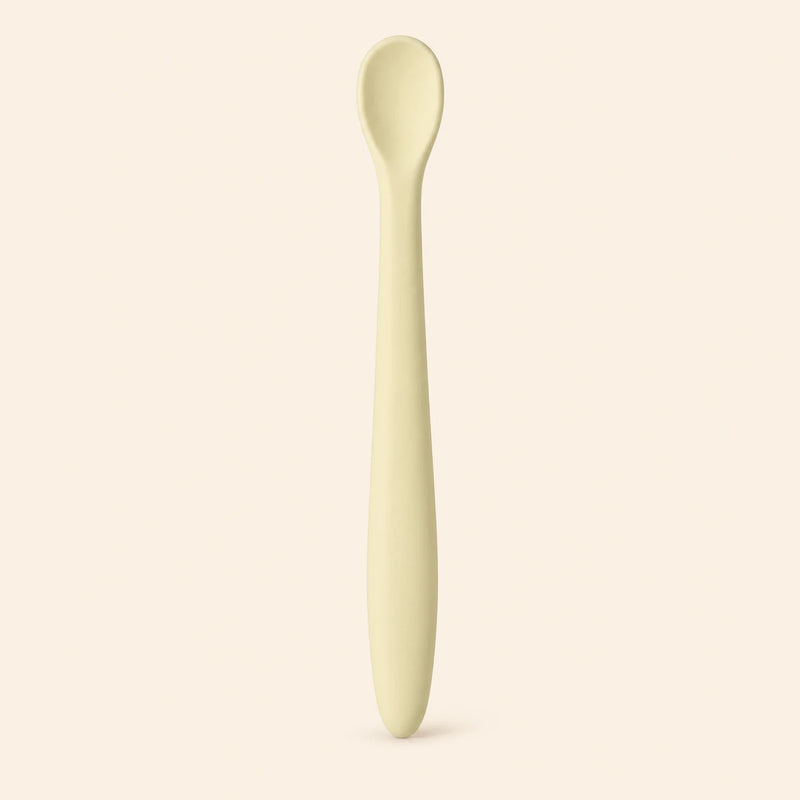 Oat silicone baby feeding spoon, showing its slim handle and shallow oval bowl on warm beige.