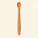 Caramel silicone baby feeding spoon, showing its slim handle and shallow oval bowl on warm beige.