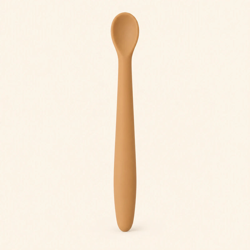 Caramel silicone baby feeding spoon, showing its slim handle and shallow oval bowl on warm beige.
