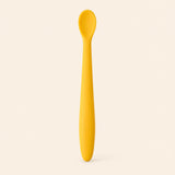 Honey silicone baby feeding spoon, showing its slim handle and shallow oval bowl on warm beige.