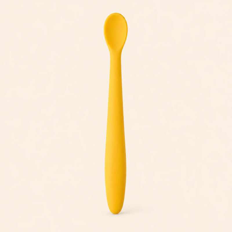 Honey silicone baby feeding spoon, showing its slim handle and shallow oval bowl on warm beige.