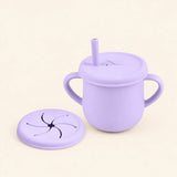 Lilac silicone cup with two side handles, fitted straw lid and straw, plus a separate snack lid on warm beige.