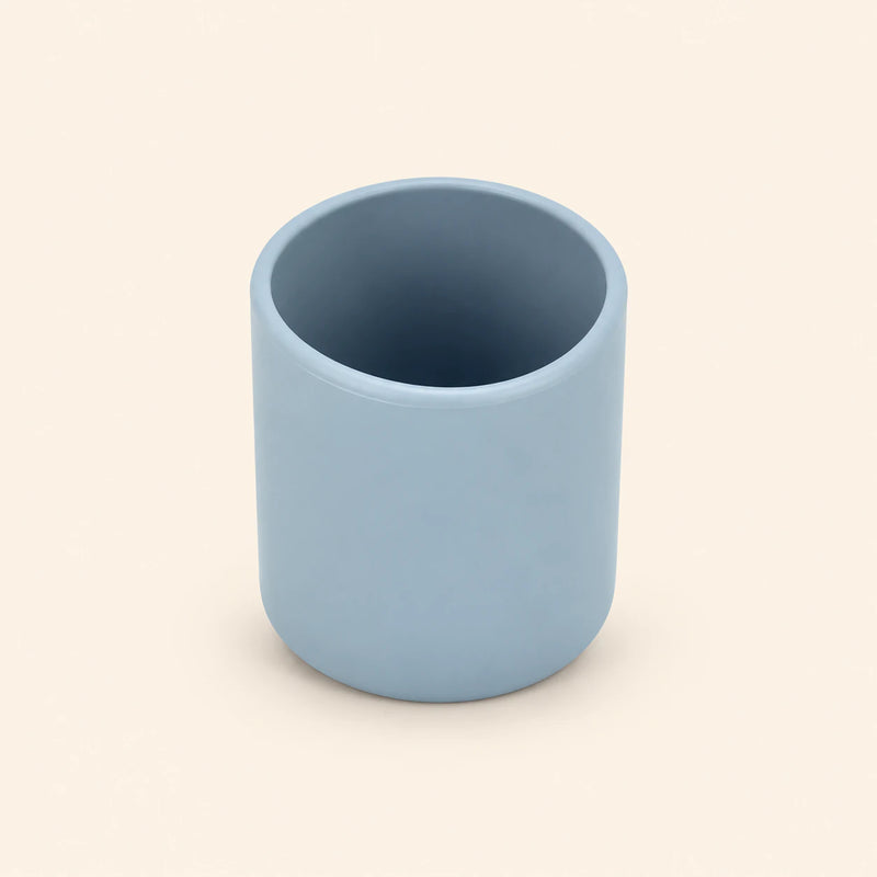Grey blue open silicone training cup with a smooth rounded rim on warm beige.