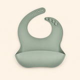 Sage Green silicone catch bib, with a scoop pocket and adjustable button-and-hole neck closure, on warm beige.