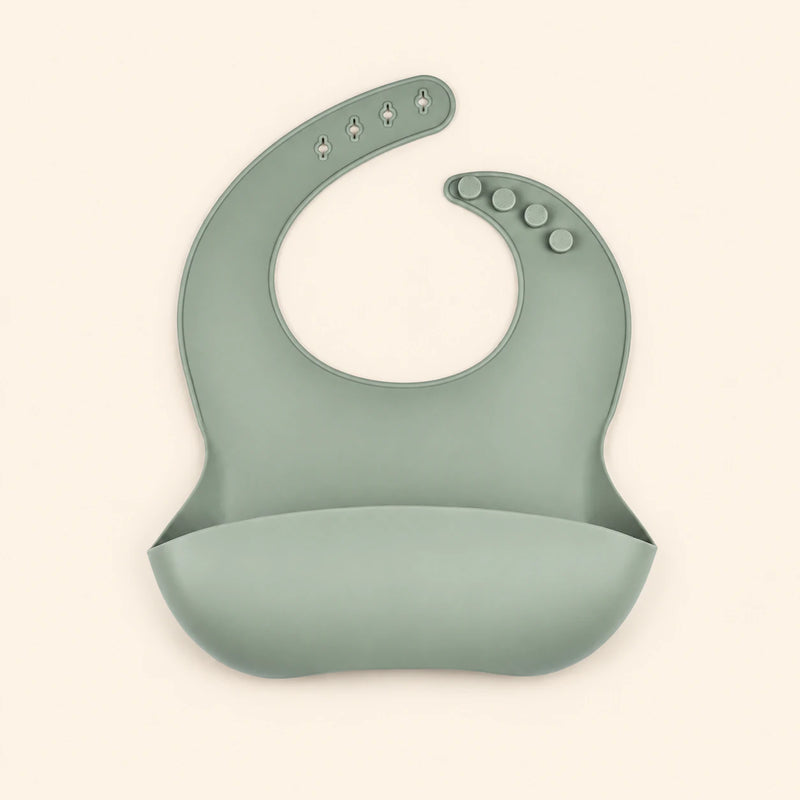 Sage Green silicone catch bib, with a scoop pocket and adjustable button-and-hole neck closure, on warm beige.
